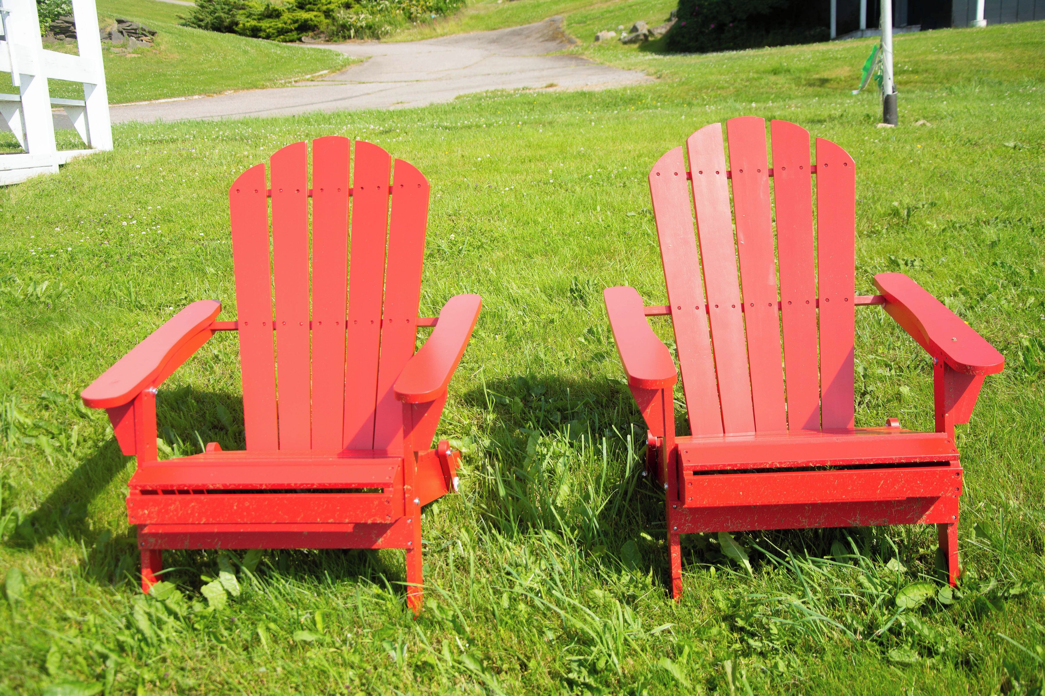Two red Adirondack chairs in a grassy area with a white fence and a paved path in the background.