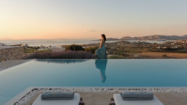A woman standing by an infinity pool with a view of the sea and hills. - H2O Private Villa Paros, infinity swimming pool, sea view lounge area, bbq & Bar (Koukoulavmes)