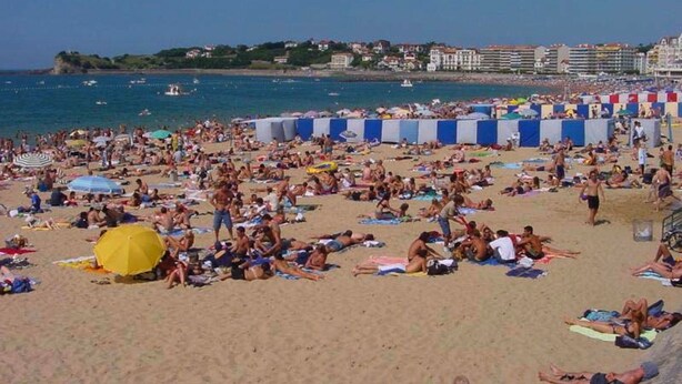 Una playa concurrida, gente tomando el sol y nadando, cielo azul despejado y edificios al fondo.