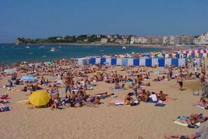 Une plage bondée où les gens se prélassent au soleil et se baignent, un ciel bleu dégagé et des bâtiments en arrière-plan.