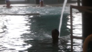 Una piscina interior con agua cristalina, una fuente visible y una persona nadando.