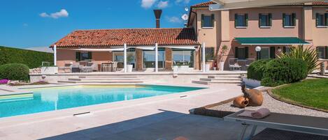 A swimming pool with a pergola overhead, a covered patio area, and a building with a tiled roof and multiple windows.