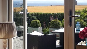 Uitzicht vanuit een kamer met een glazen deur, op een aangelegde tuin met een haag, een tafeltje met een lamp en een plant in pot.