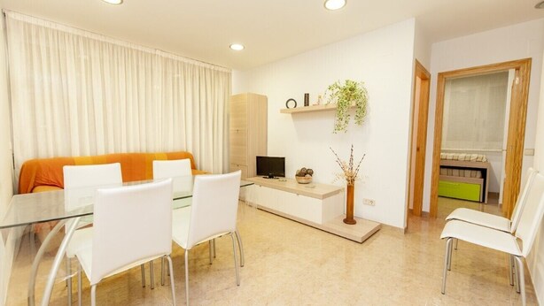 A modern living room with a glass dining table, white chairs, a sofa with orange cushions, a TV, a plant, and a window with a view. - Hermitage 1stA (Peñiscola)