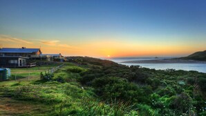 A coastal landscape with a house overlooking the sea, a grassy area with wildflowers, and a sunset. - Arthur River Spa Cottage (Arthur River)