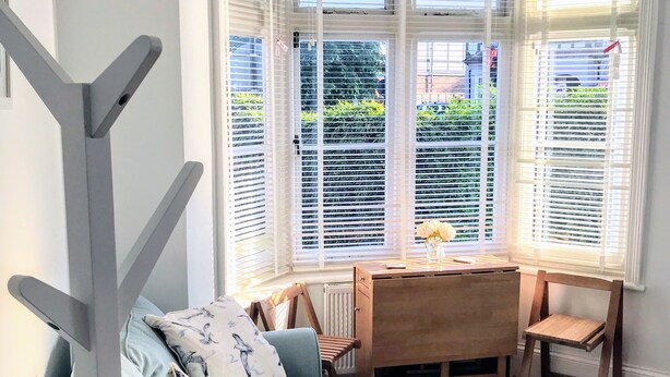 Un salón moderno con un sofá gris, una mesa de comedor de madera y sillas. Un ventanal con persianas blancas ofrece vistas a la vegetación exterior.