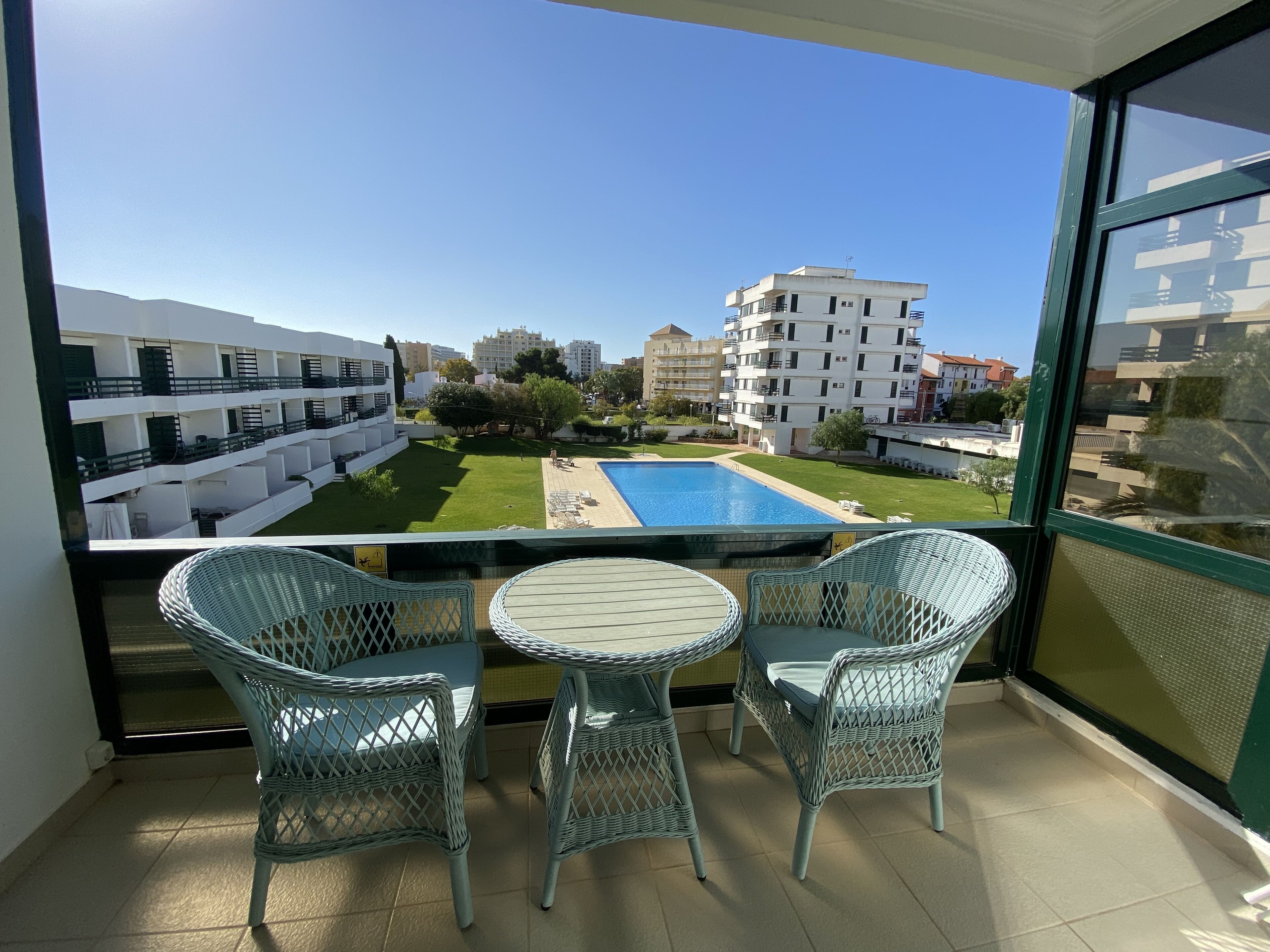 Un balcone con sedie in vimini e un tavolino, con vista su una piscina e un cortile con edifici sullo sfondo.