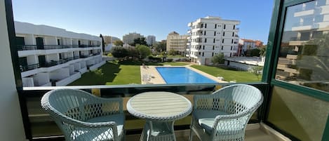 Un balcón con sillones de mimbre y una pequeña mesa, con vistas a una piscina y un patio con edificios al fondo.