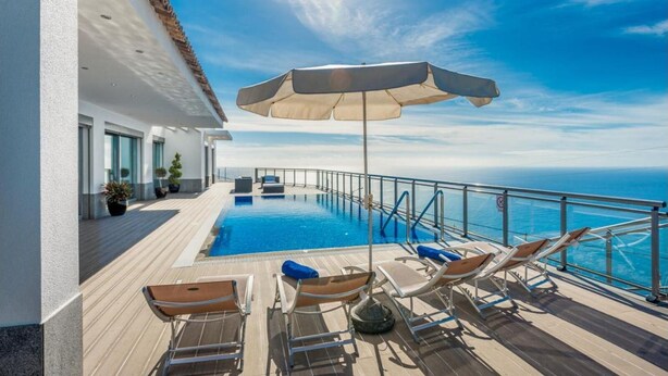 A sunlit outdoor swimming pool with lounge chairs and umbrellas, overlooking the sea.