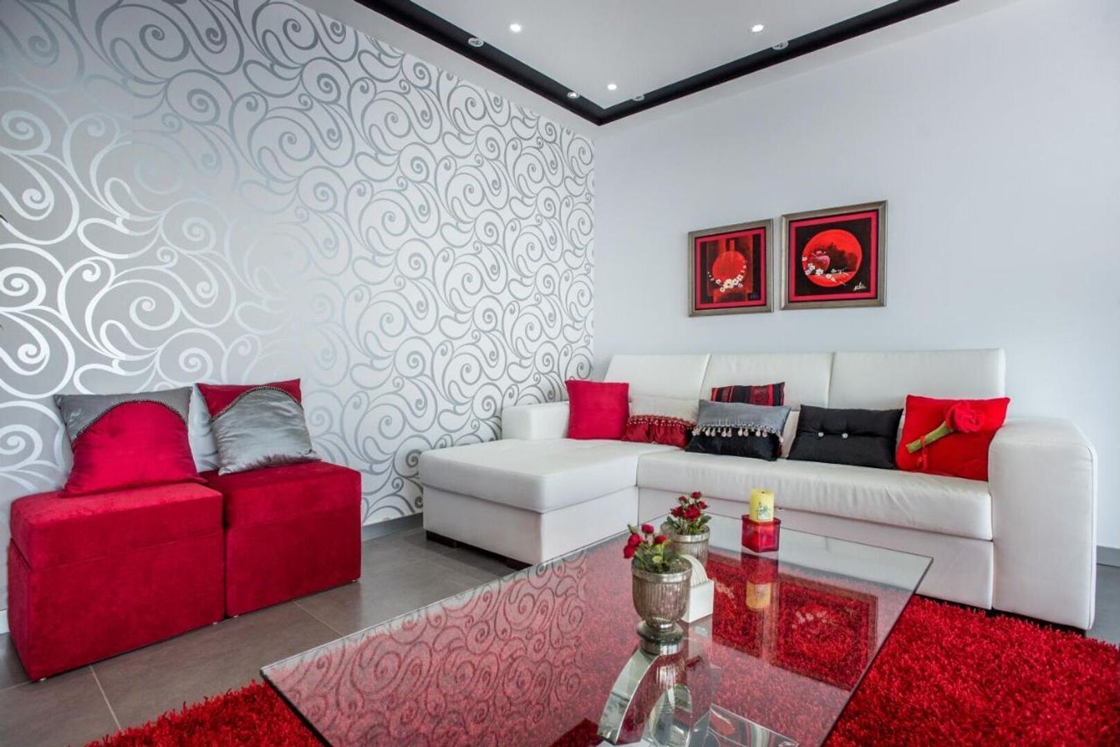 Un salon moderne avec un canapé blanc, des coussins rouges et gris, une table basse en verre et un papier peint à motifs.