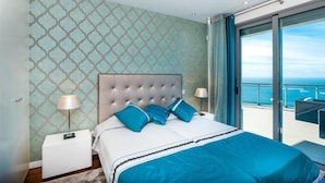 A bedroom with a large bed, two bedside lamps, a nightstand, and a view of the ocean through a balcony.
