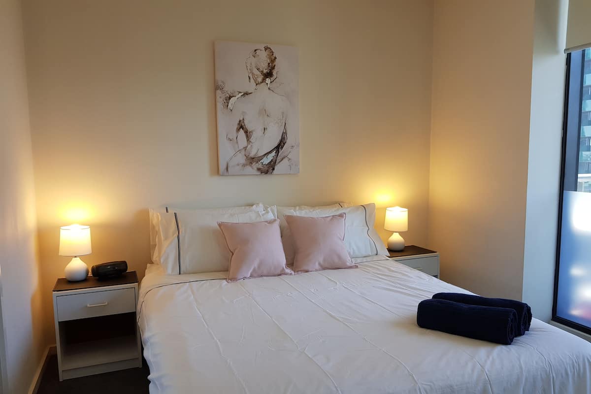 A neatly made bed with white bedding and pink pillows, two bedside lamps, a nightstand with a drawer, and a painting on the wall.
