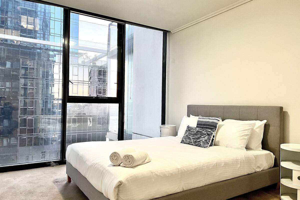 A modern hotel room with a large window, a bed with white linens, and a grey headboard.