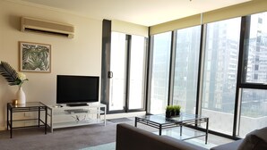 Ein modernes Wohnzimmer mit großem Fenster, einem Flachbildfernseher, einer Couch, einem Couchtisch und einem kleinen Beistelltisch mit Vase und Dekoration.