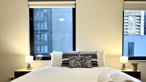 Una cama bien hecha con sábanas blancas y toallas enrolladas, dos lámparas de noche y una ventana con vista a un edificio.