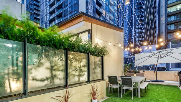 Un patio moderno al aire libre con comedor, césped artificial, plantas en macetas y vista a un edificio de gran altura.