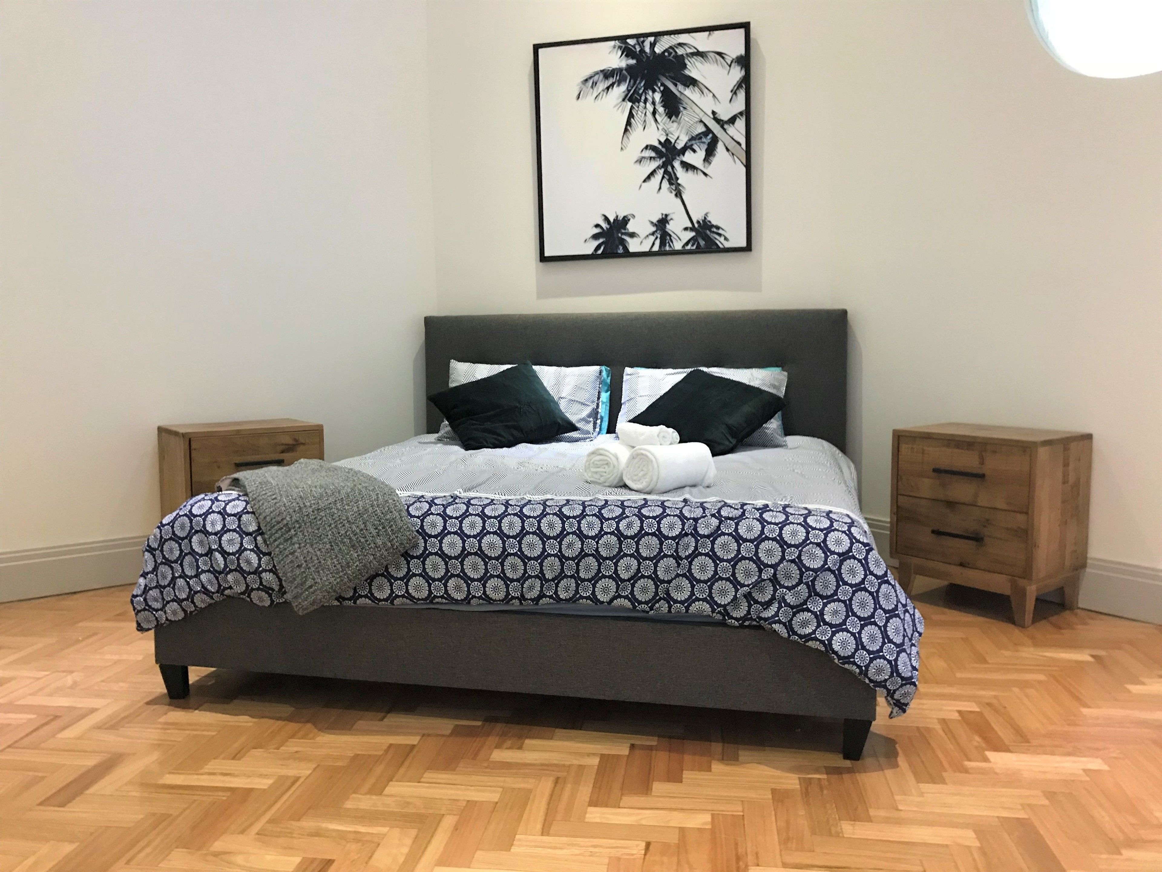 Una cama bien hecha con ropa de cama estampada, dos mesitas de noche, un cuadro colgado en la pared y un suelo de madera con diseño de espiga.