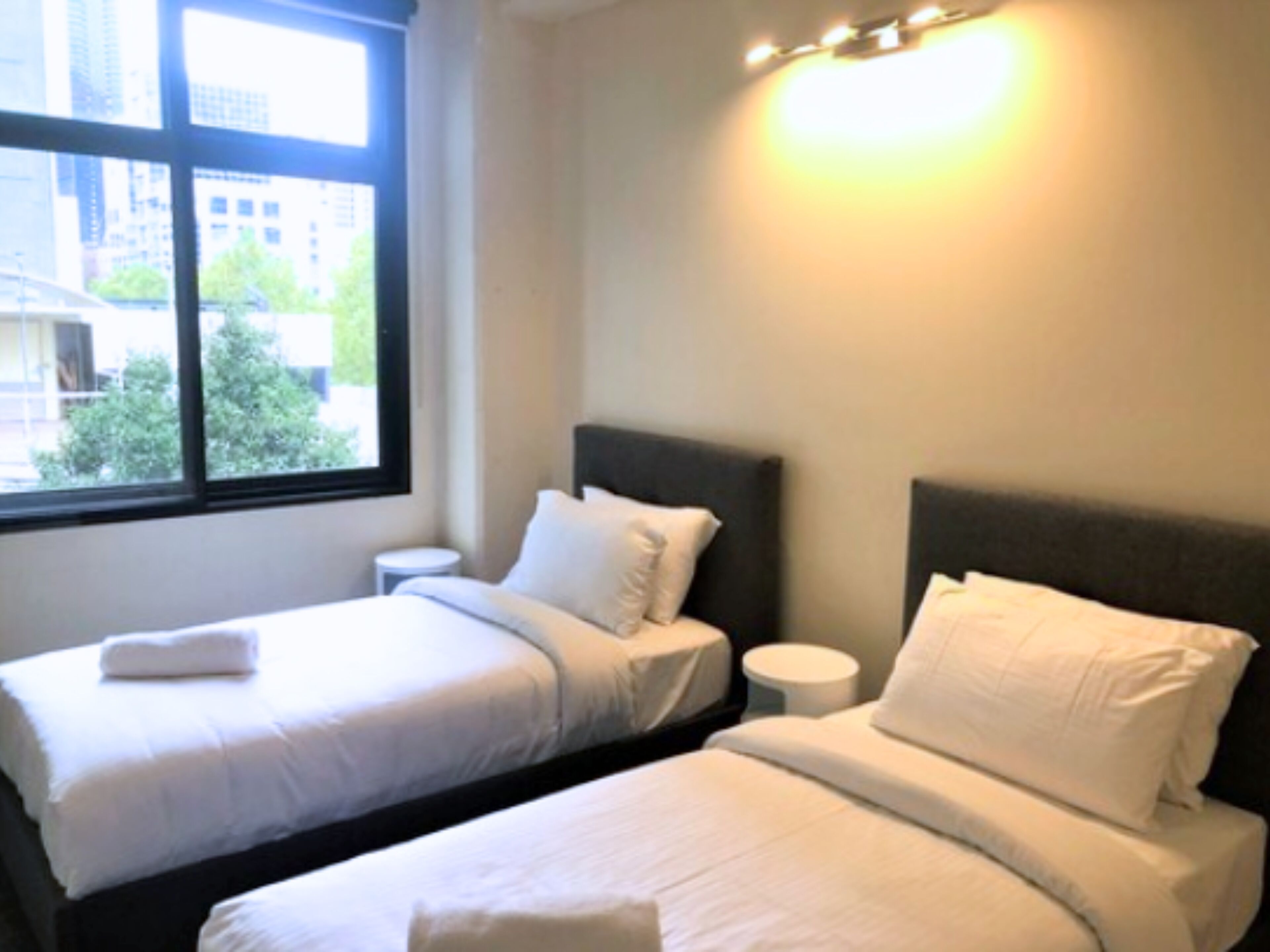 A hotel room with two beds, a window with a view of buildings and trees, and a bedside table with a lamp.