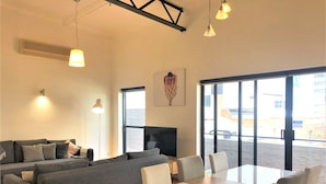 Una sala moderna con un sofá seccional, una mesa de comedor con sillas, lámparas colgantes y ventanas amplias que ofrecen vistas al exterior.