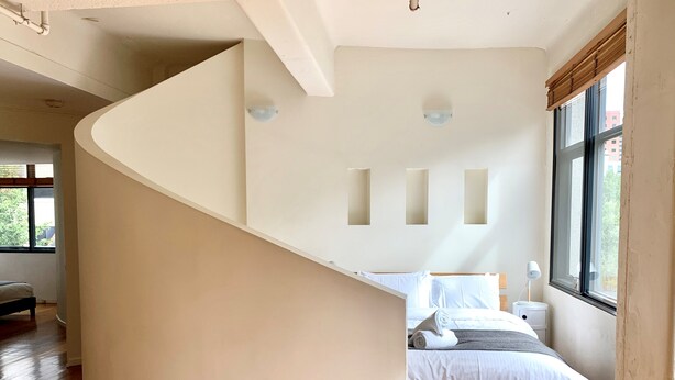 Un dormitorio moderno con una escalera curva, una cama con ropa de cama blanca y un ventanal con vistas a la vegetación.