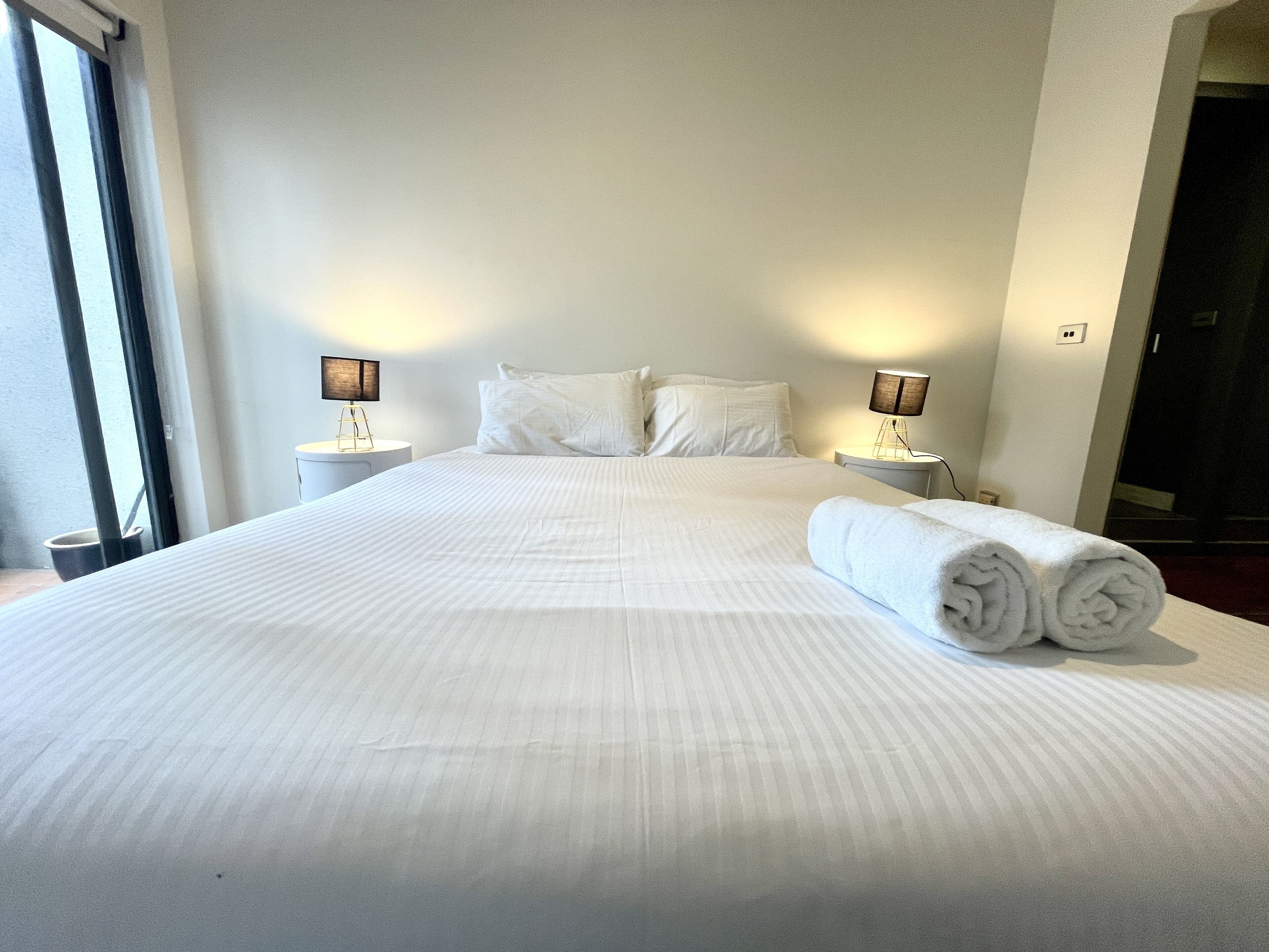 Una cama grande con ropa blanca, dos mesitas de noche con lámparas y una toalla enrollada sobre la cama.
