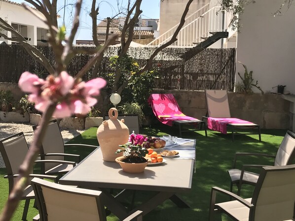 Un patio al aire libre con una mesa lista para una comida, rodeada de sillas. Hay plantas en macetas sobre la mesa, y una silla rosa es visible al fondo.