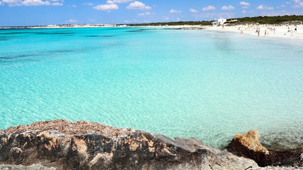 A beach with clear turquoise water, rocky foreground, and a distant shoreline with buildings.