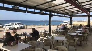 An outdoor cafe with tables and chairs, overlooking the sea and a road with vehicles.