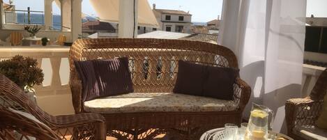 A balcony with wicker furniture, a glass table with yellow drinks, and a view of the sea and buildings.