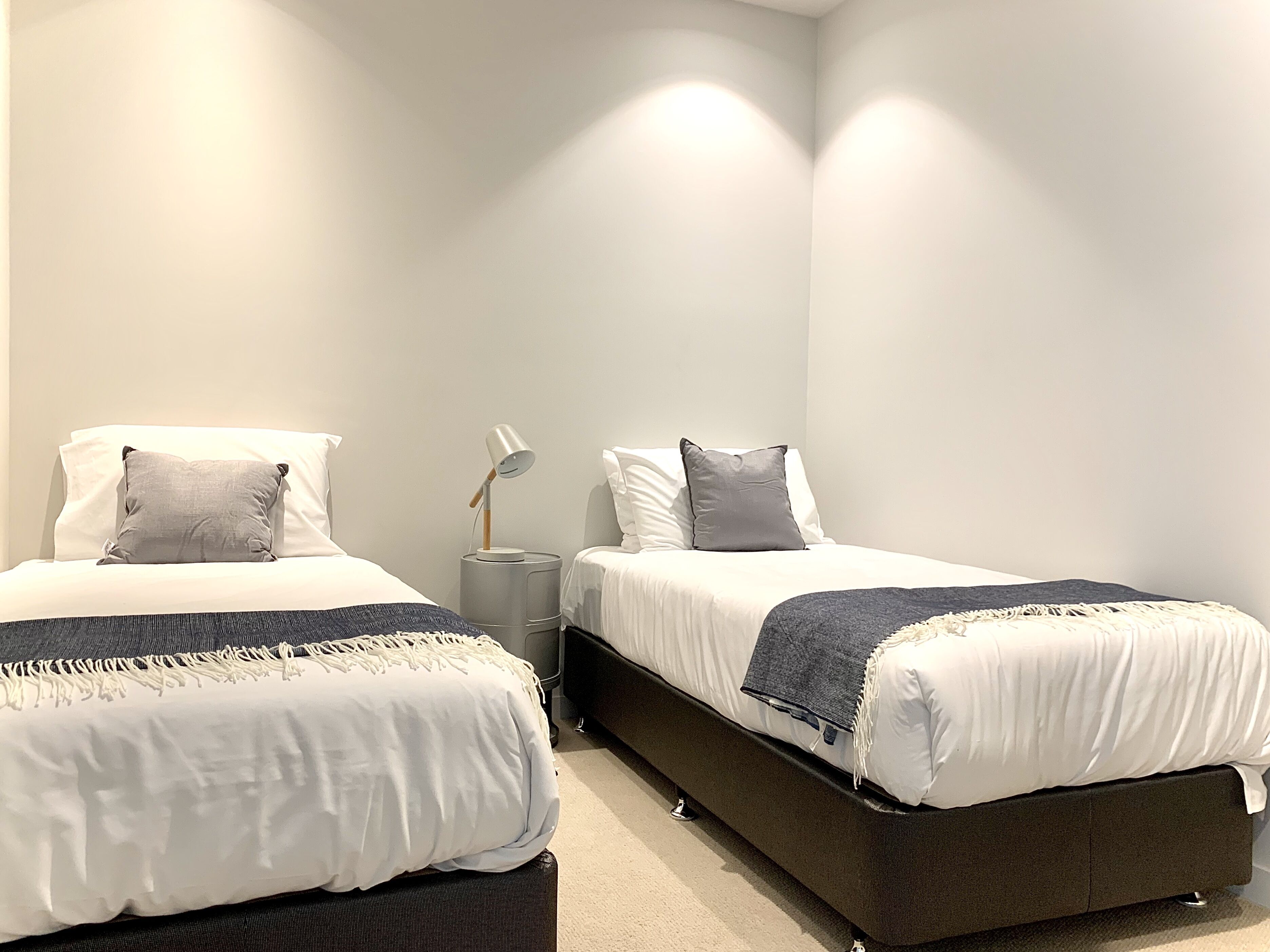 A bedroom with two beds, each with white linens and grey pillows, a grey nightstand with a lamp, and a dark-colored bench at the foot of the beds.