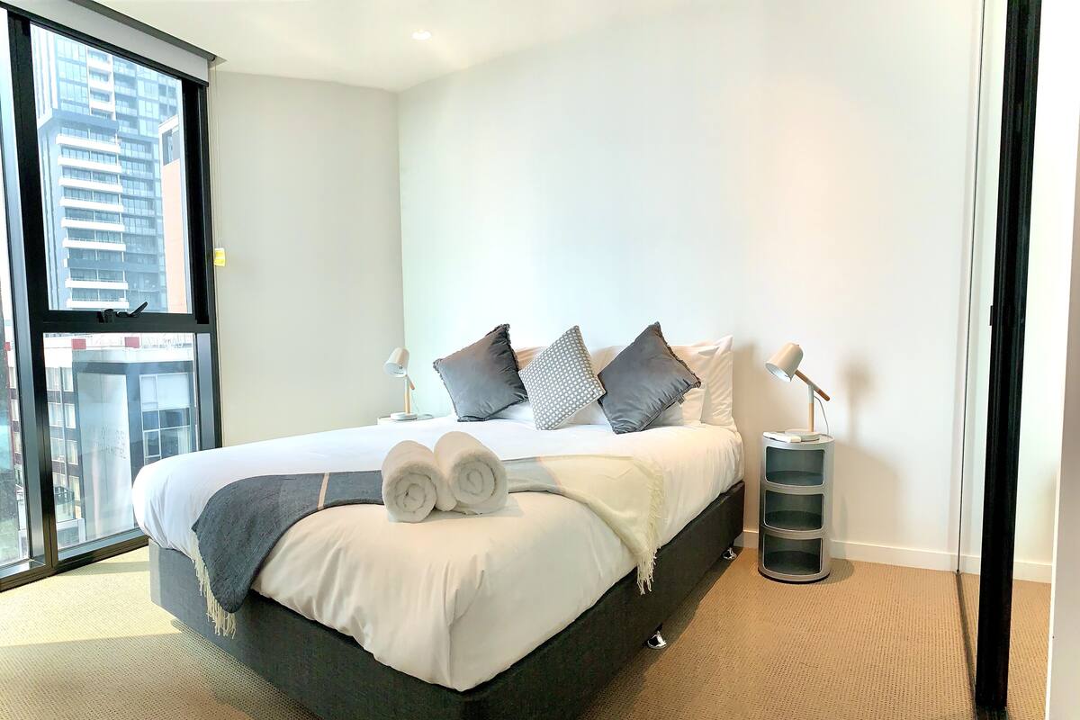 Una habitación de hotel moderna con una cama grande, dos lámparas de noche, una mesita auxiliar y vistas a un paisaje urbano a través de un ventanal amplio.