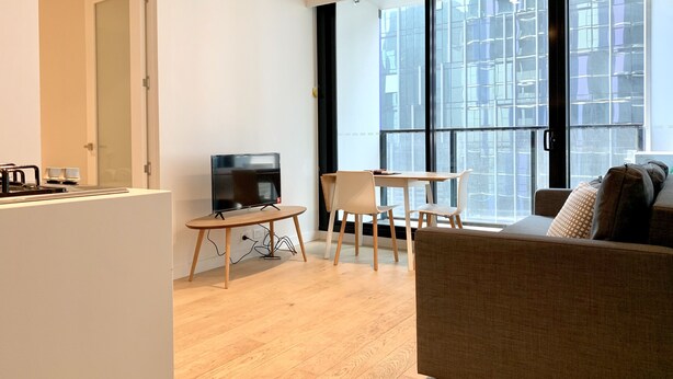 Una sala de estar moderna con un sofá, un televisor sobre un mueble bajo, una pequeña mesa con sillas y un ventanal con vistas a la ciudad.