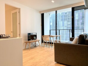 A modern living room with a sofa, a TV on a stand, a small table with chairs, and a large window with a city view. - ReadySet Apartments at Lighthouse (Melbourne)