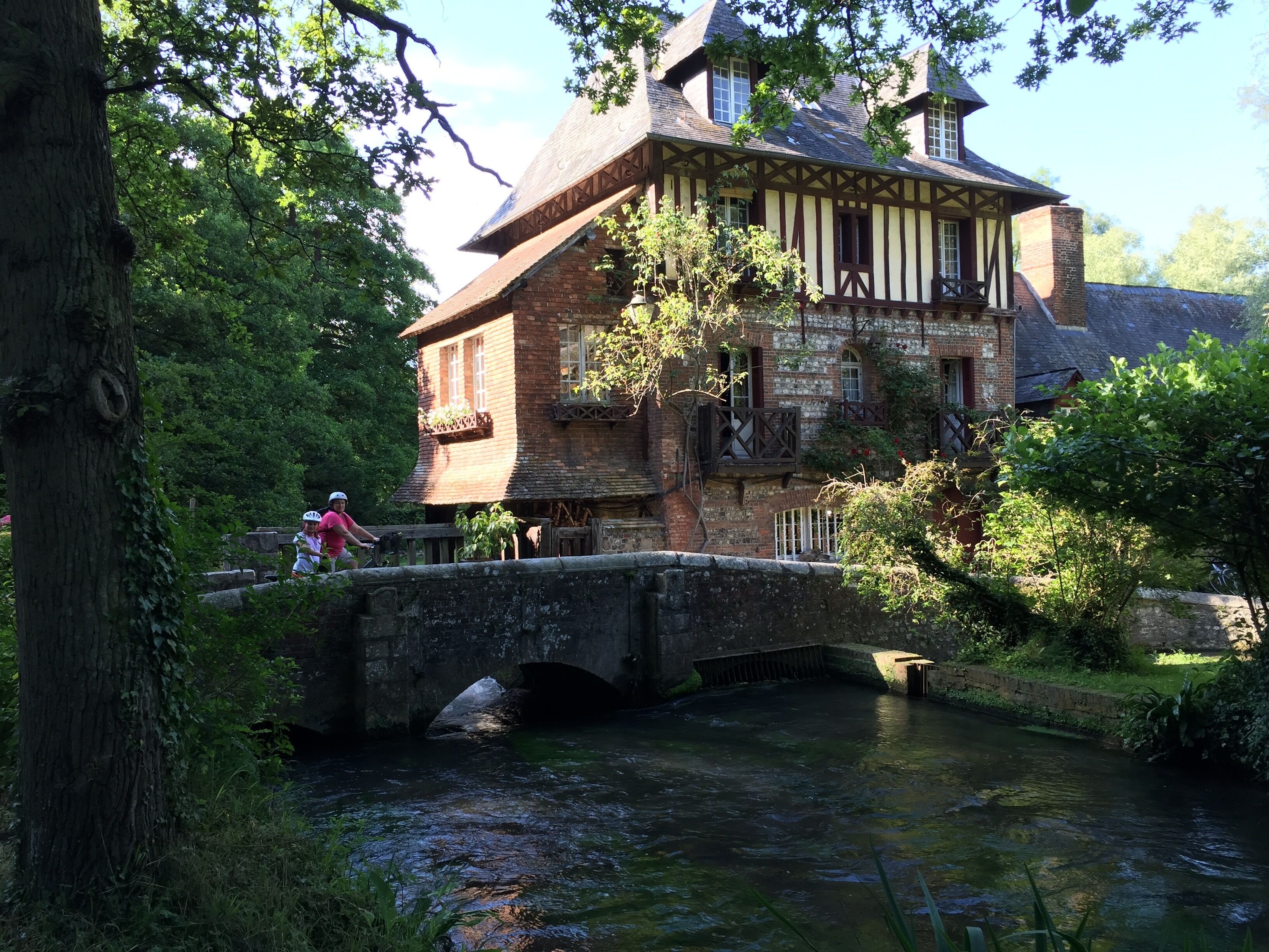 Ein historisches Gebäude im Tudor-Stil mit einer Steinbrücke über einem Fluss, umgeben von üppigem Grün.