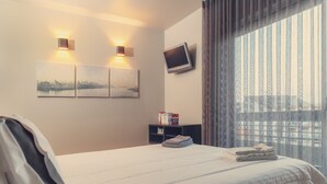 A hotel room with a neatly made bed, a flat-screen TV mounted on the wall, and a window with vertical blinds. - Porto RiverView Flat (Vila Nova de Gaia)