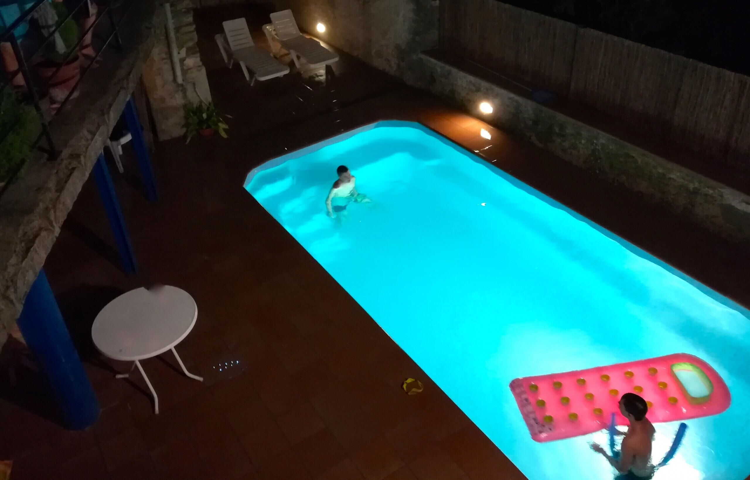 Une piscine extérieure avec une personne dans l’eau, un flotteur gonflable rose et une table et des chaises blanches à proximité.