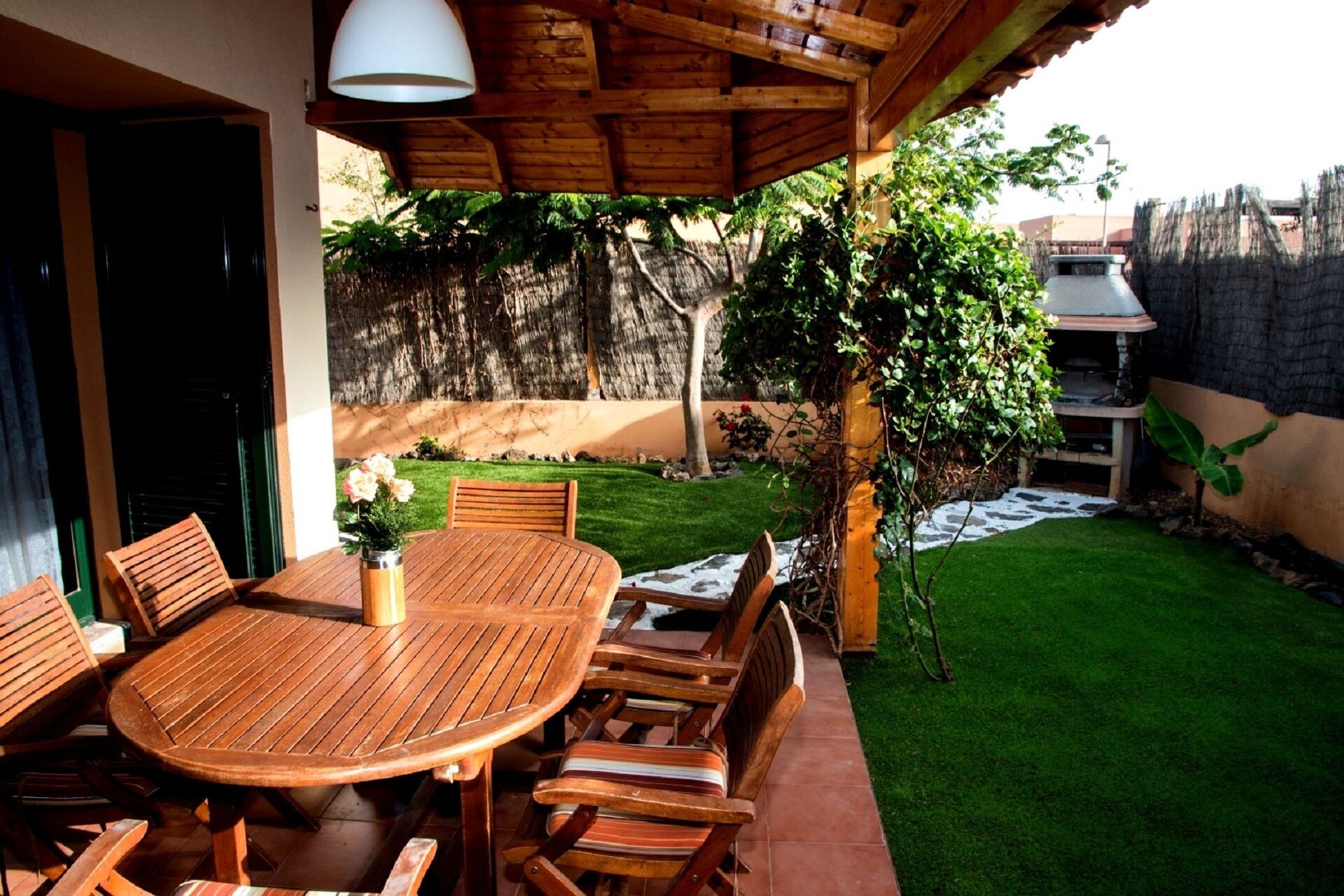 A wooden patio with a round table and chairs, a potted plant, and a thatched roof structure.
