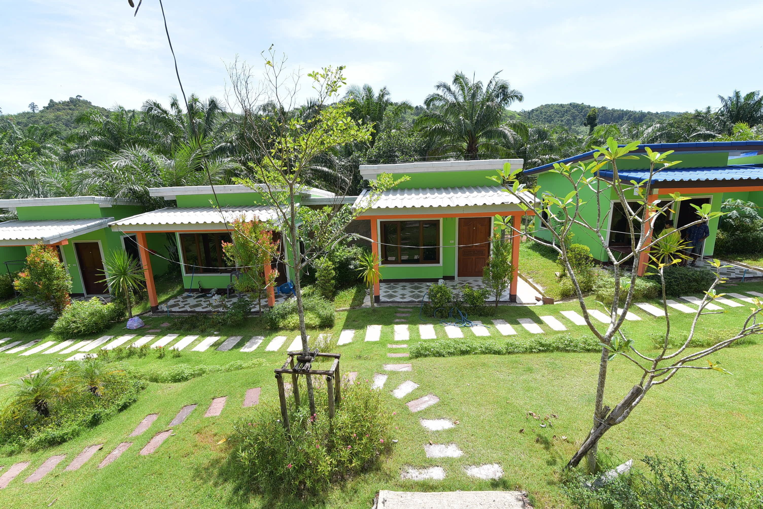 A row of green buildings with tiled roofs, surrounded by a well-maintained garden with stone pathways and lush greenery.