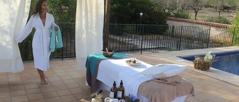 A spa area with a massage table, towels, and bottles, overlooking a pool and garden.