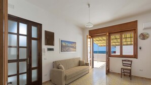 A room with a sofa, a chair, a painting, a clock, and a view of the sea through a window. - Appartamento Orizzonte by BarbarHouse (Porto Cesareo)