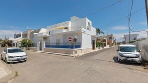 A white building with a blue balcony, a no entry sign, and two parked cars on a clear street. - Appartamento Orizzonte by BarbarHouse (Porto Cesareo)