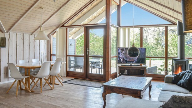 Een ruime woonkamer met een houten plafond, grote ramen, een televisie, een open haard en een eethoek met stoelen en een tafel.
