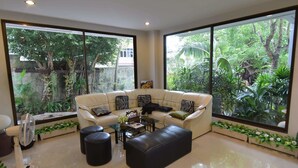 A spacious living room with a large sofa, black ottoman, and a coffee table. There are two large windows offering a view of lush greenery. - Kyongean Mansion 2 (Krabi)