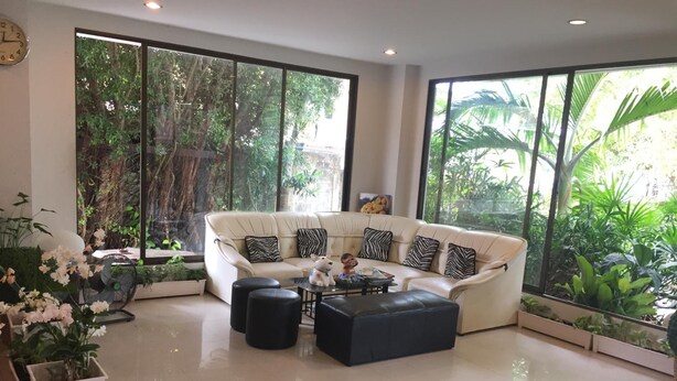 A modern living room with a white sofa, black and white patterned cushions, a coffee table, and a decorative dog figurine. Large windows provide views of lush greenery outside. - Kyongean Mansion 2 (Krabi)