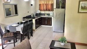 Cocina compacta con refrigerador, microondas y lavamanos. Hay un comedor con mesa y sillas, y una pequeña sala de estar con sofá y mesa de centro.