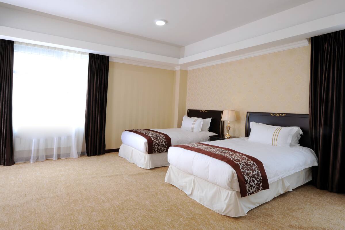 A hotel room with two beds, a large window with curtains, a nightstand with a lamp, and a headboard with a decorative design.