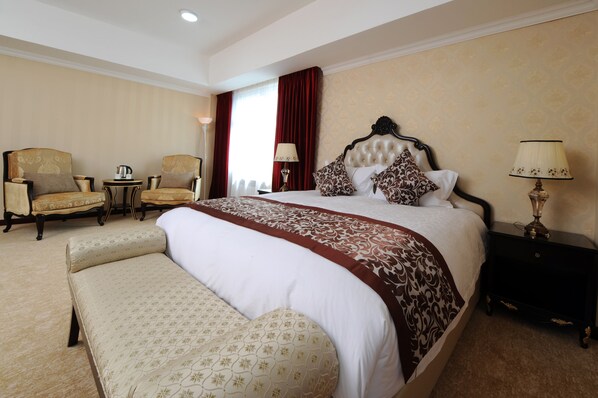 Una habitación de hotel con una cama grande, dos sillones, una mesita de noche con lámpara y un banco.