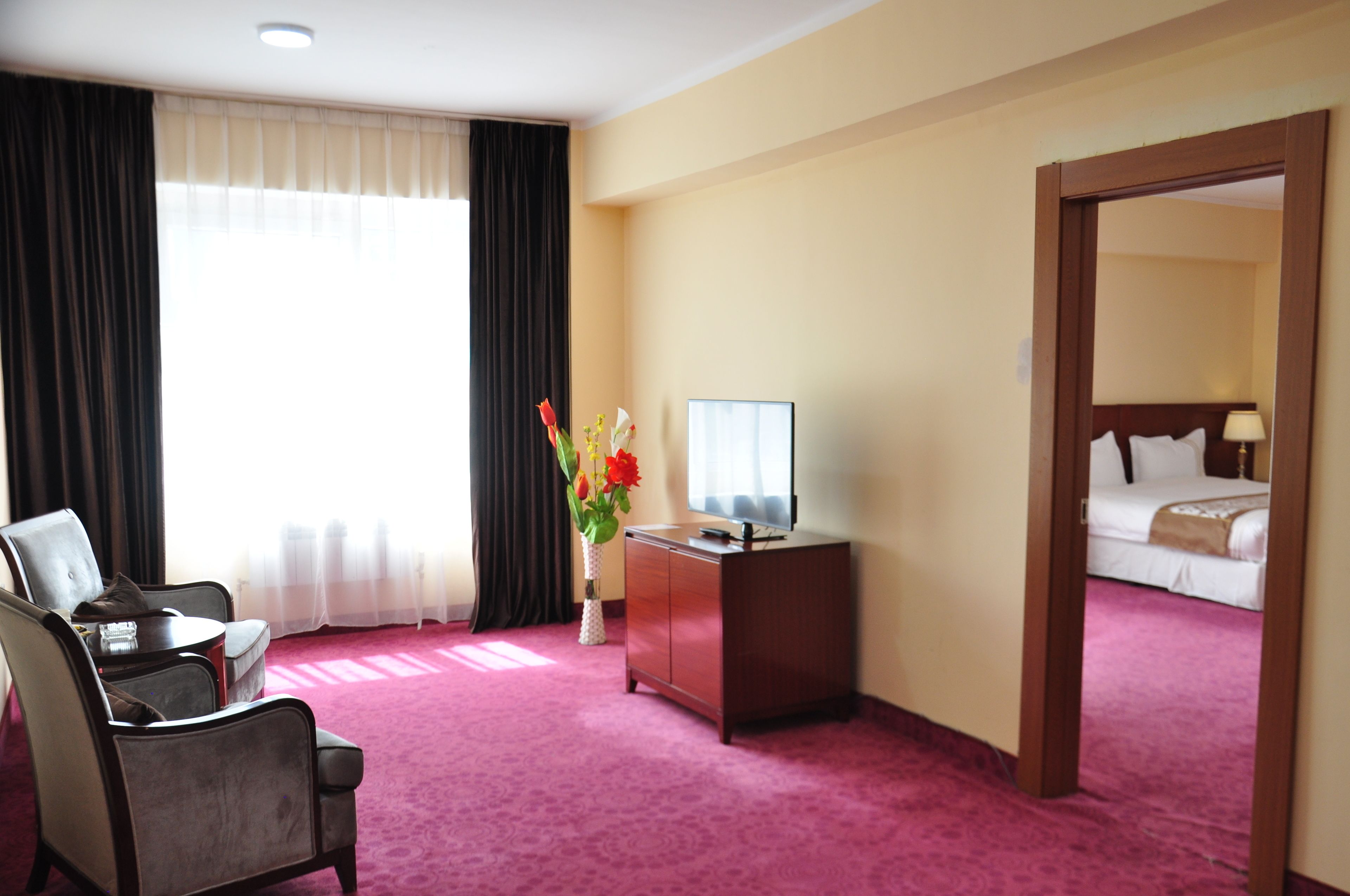 Hotelkamer met een bed, een bureau met computer, een stoel, een vaas met bloemen en een groot raam met gordijnen.