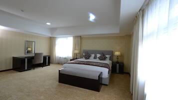 A hotel room with a large bed, a desk, a chair, a bedside table with a lamp, and a window with curtains.