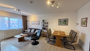 Un salon moderne avec un canapé, des fauteuils, une table basse et une table à manger. Les murs sont ornés de tableaux encadrés et une plante est posée sur une étagère.